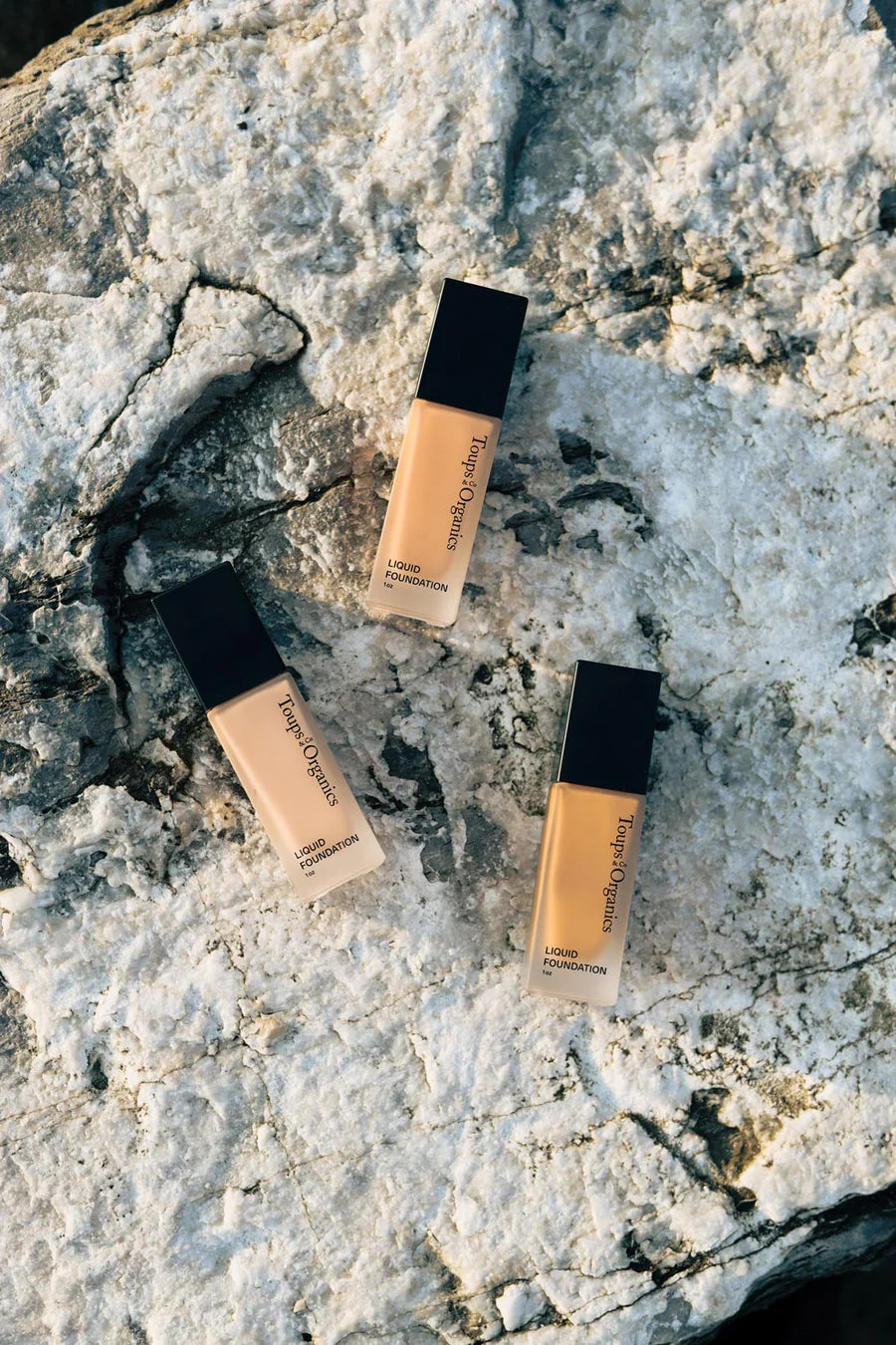 Three bottles of cosmetic foundation, in different shades, arranged on a rock.